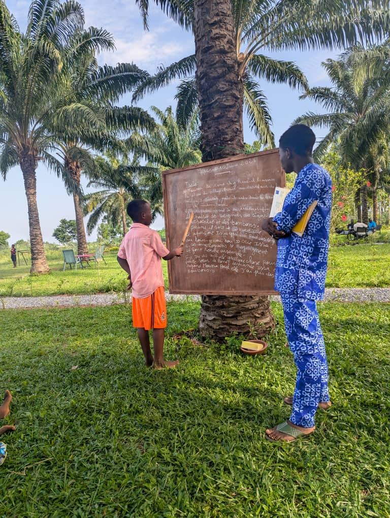 Un animateur accompagne un enfant à l'ardoise, sous les palmiers du Jardin Joseph